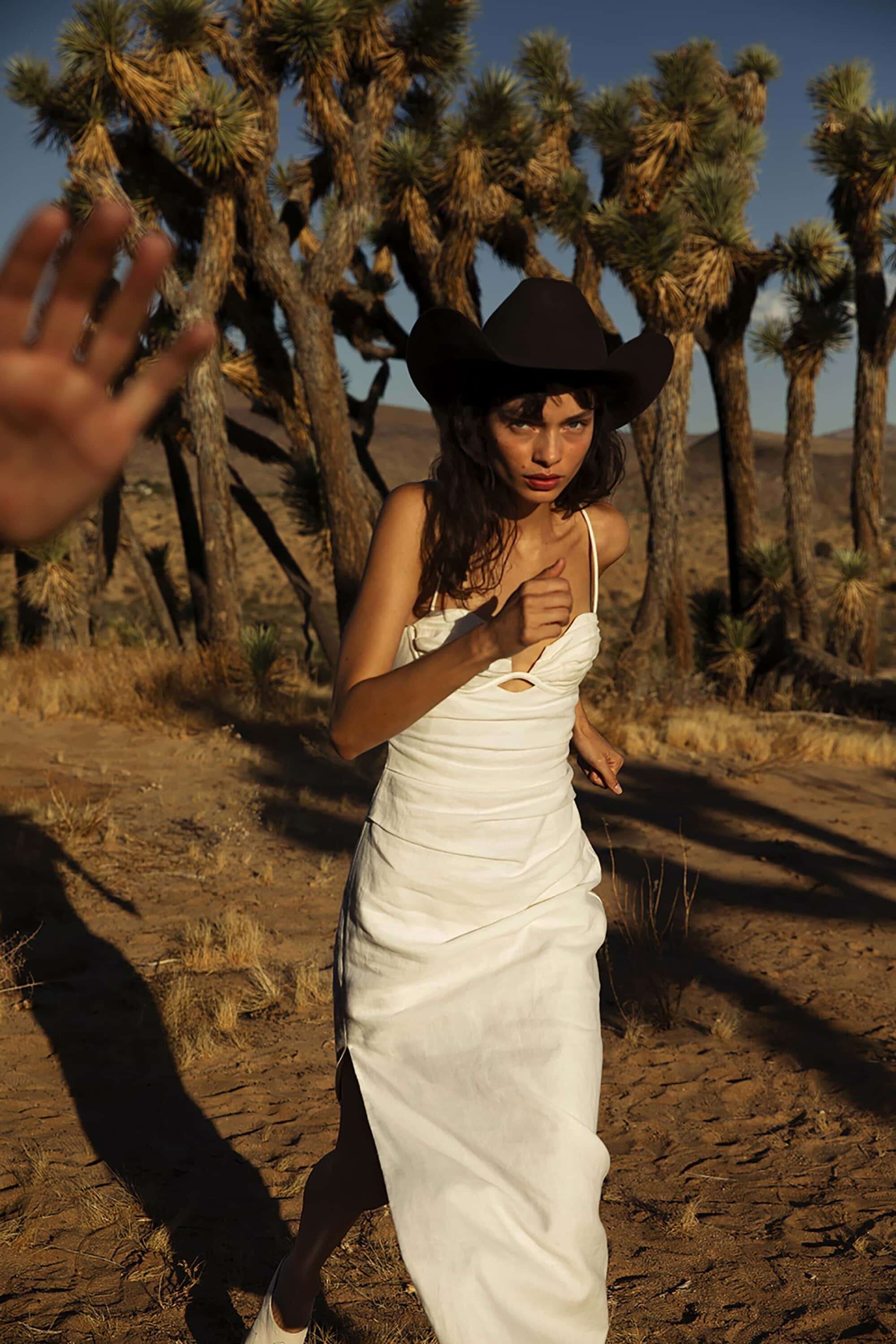 A woman in a white dress and black cowboy hat stands in the desert with Joshua trees in the background and a hand reaching in.