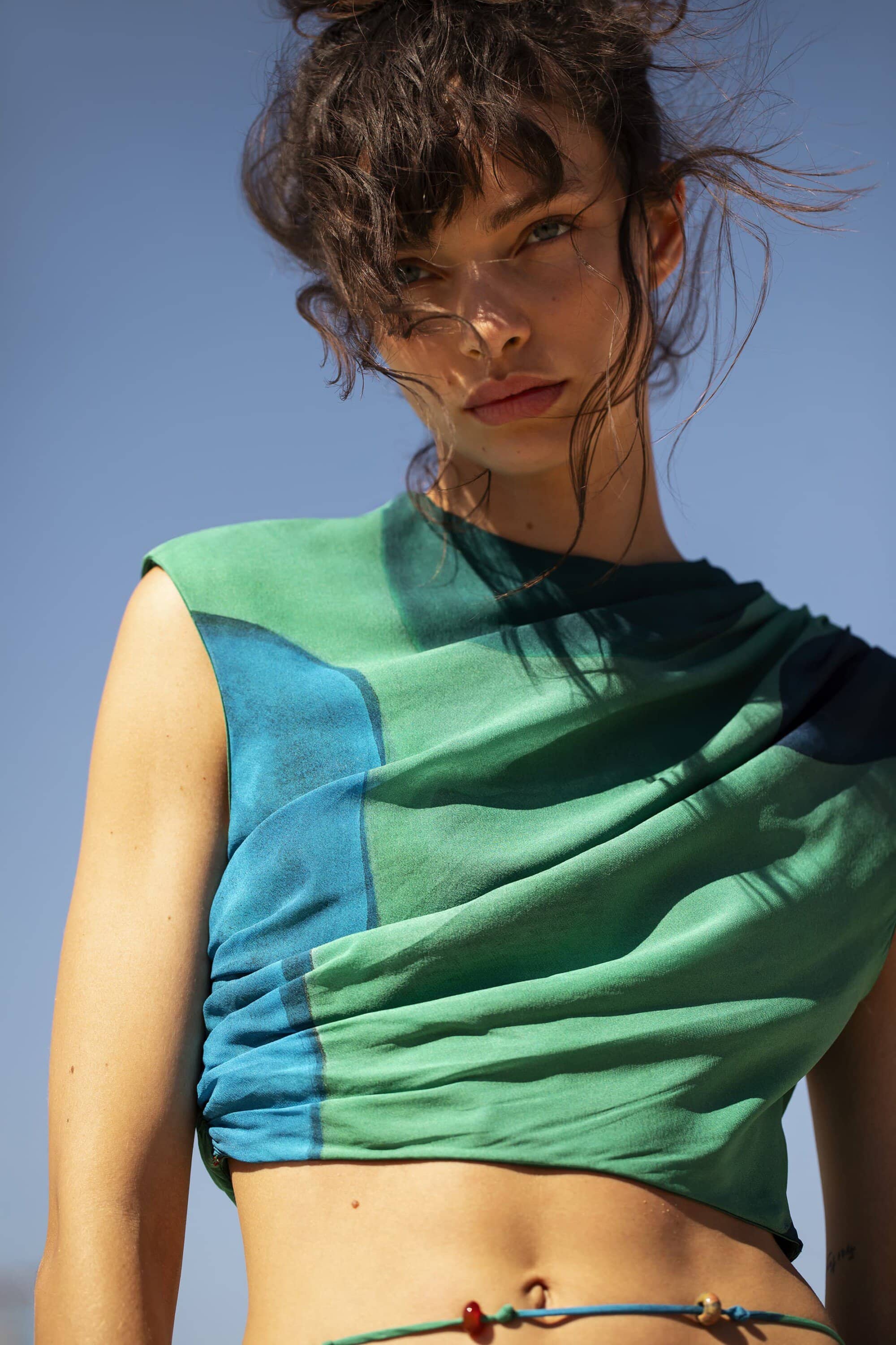 Eye-level shot of a woman with an updo hairstyle in green and blue sleeveless top against a light blue sky in outdoor setting.