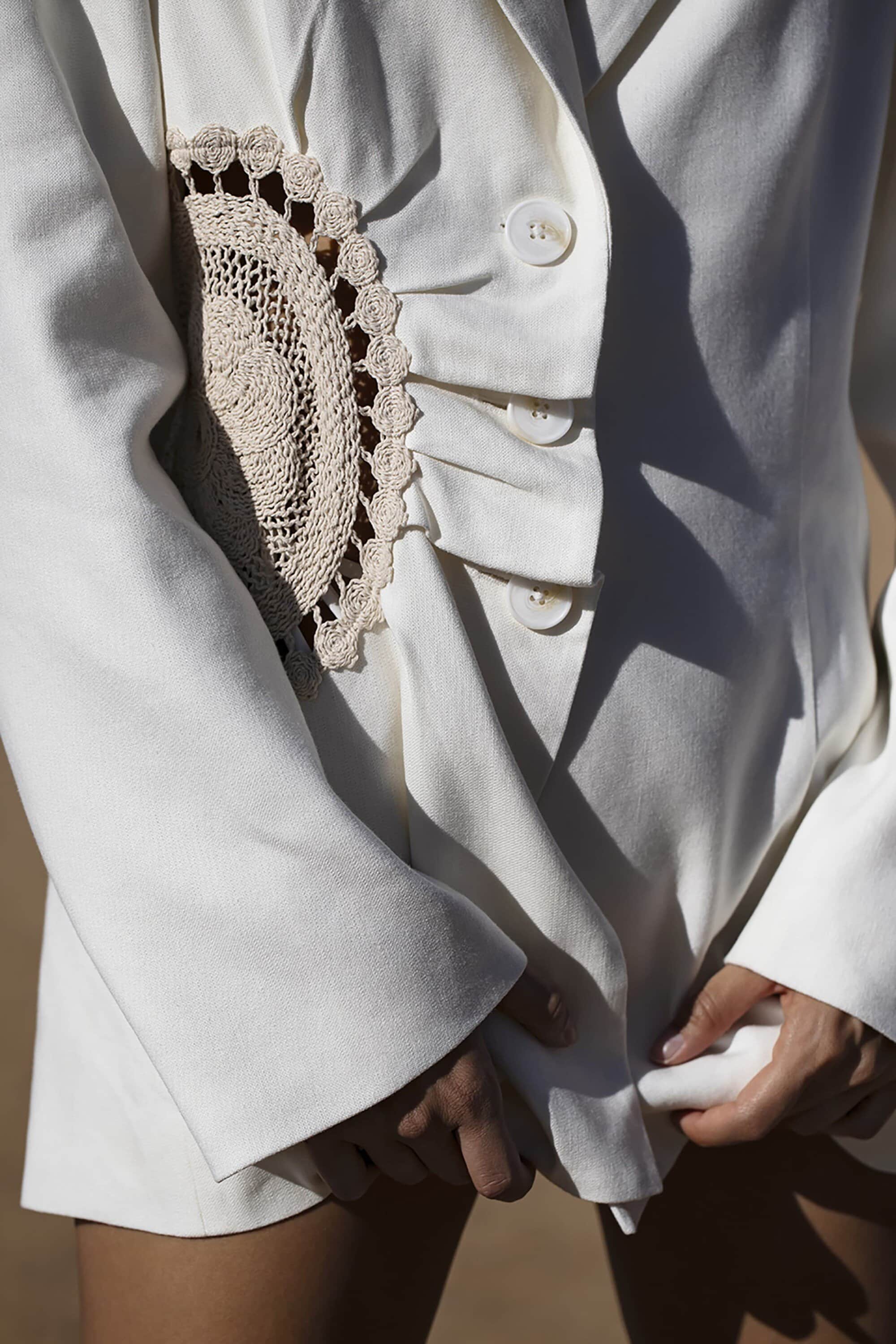 Close-up of a fair-skinned human in a white blazer with a unique crocheted patch detail. The blazer has button closures.
