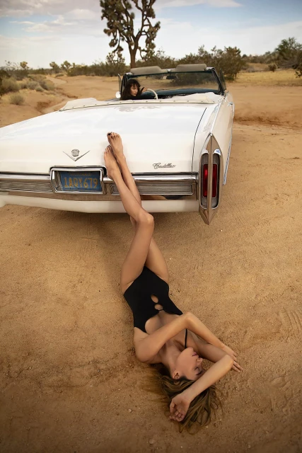 Woman in black swimsuit poses on desert ground near white Cadillac convertible, another woman in the driver's seat, Joshua trees.