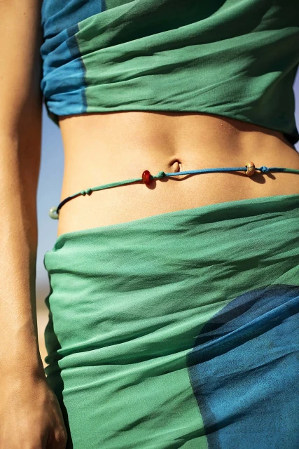 Close-up of a slender adult female torso, featuring a navel and a beaded waist string over blue and green patterned sheer fabric.