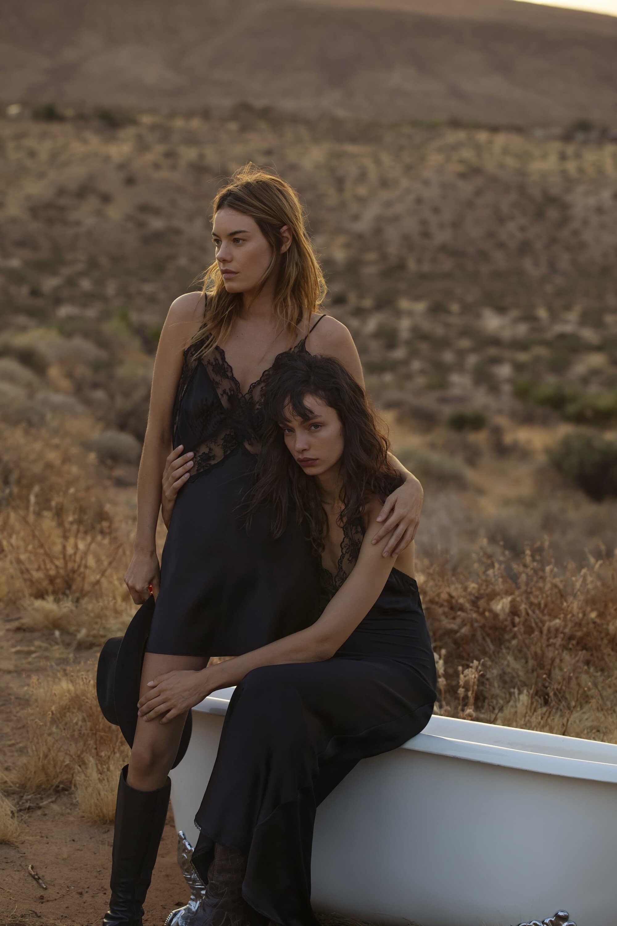 Two women in black dresses pose in a desert landscape next to a white bathtub with a hat and boots, creating a striking fashion shot.