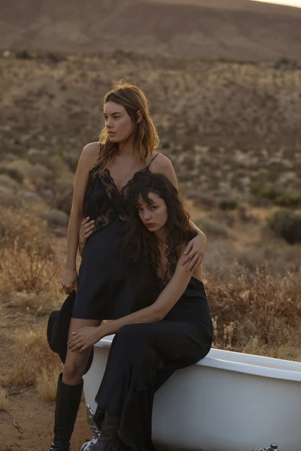 Two women in black dresses pose in a desert landscape next to a white bathtub with a hat and boots, creating a striking fashion shot.