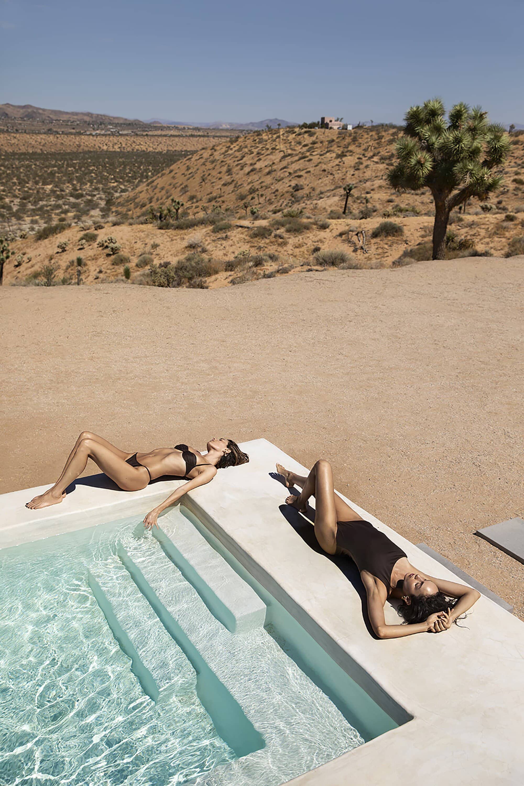 Two women in swimsuits sunbathe by a modern pool in a desert landscape with sparse vegetation and distant mountains under a clear sky.