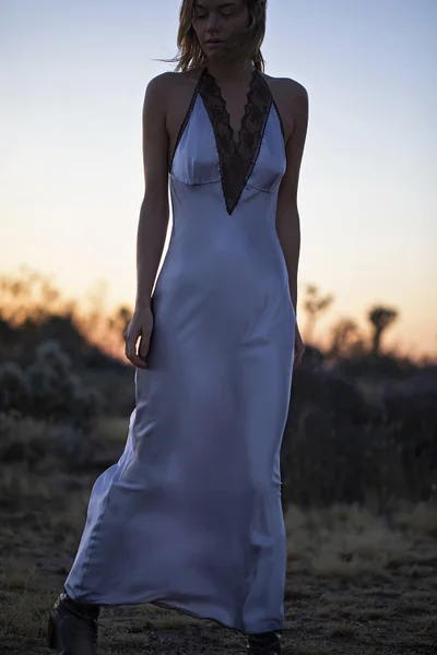 Model standing outdoors at sunset wearing a light blue satin halter dress with black lace trim.