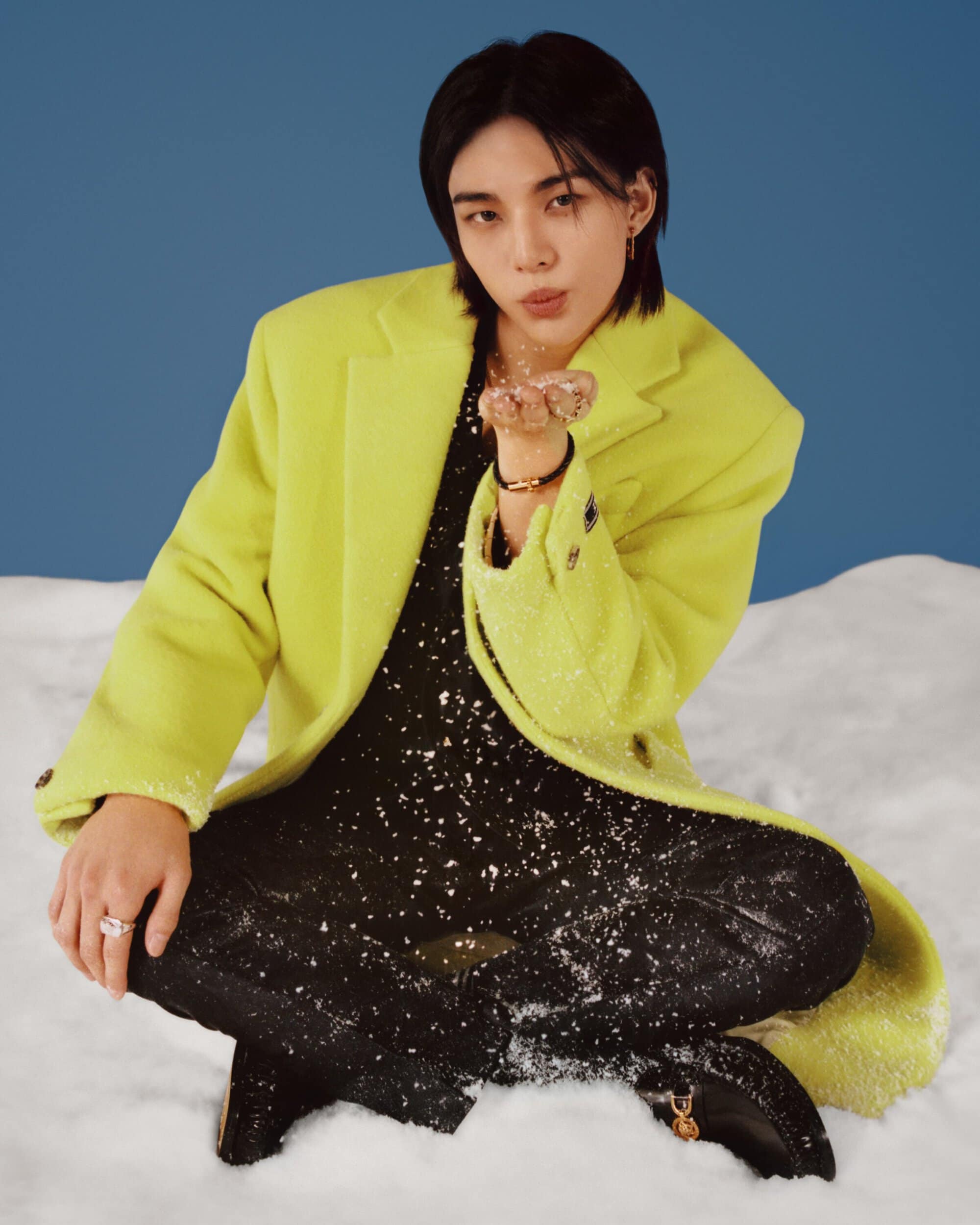 Portrait of an adult man with black hair wearing a yellow coat, squatting with his hand out, against a blue and white background.