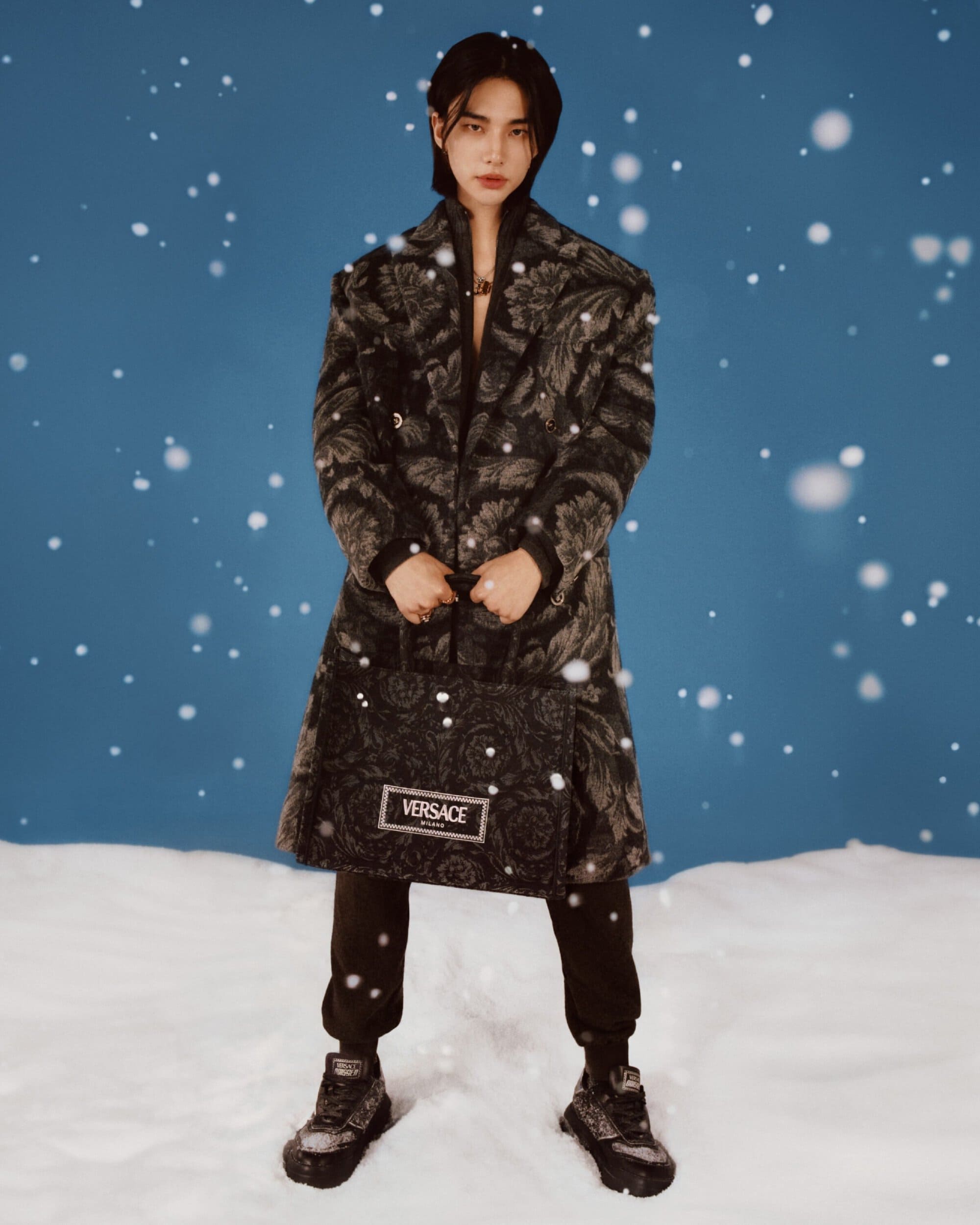 A single male stands in the snow wearing a patterned overcoat and carrying a matching Versace tote bag against a blue backdrop.