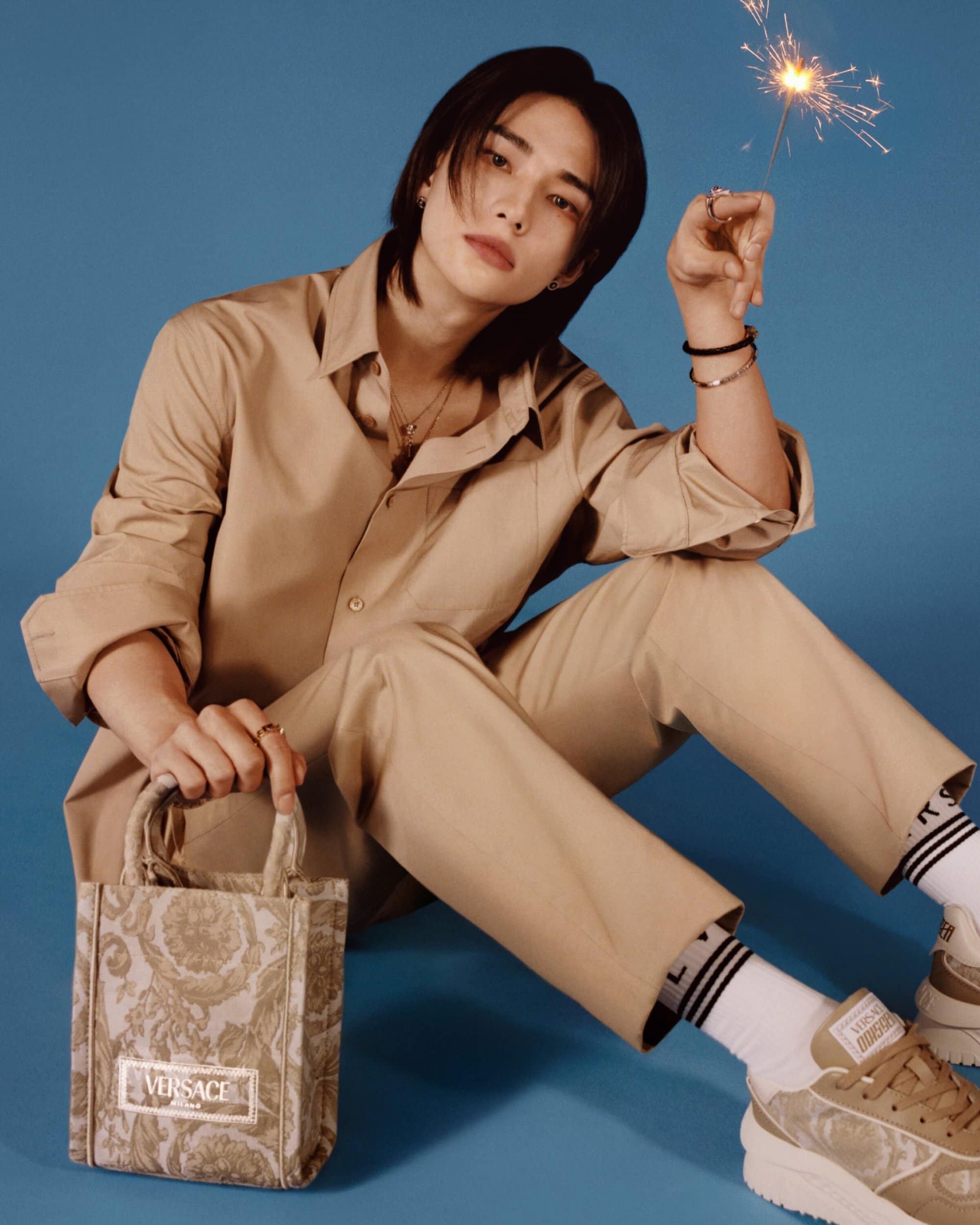 Asian male in beige outfit sits with Versace bag and sparkler against blue background in studio setting, fashion lifestyle portrait.