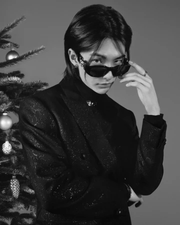 Stylish adult male poses in black suit and sunglasses next to Christmas tree in a studio setting, captured in black and white.
