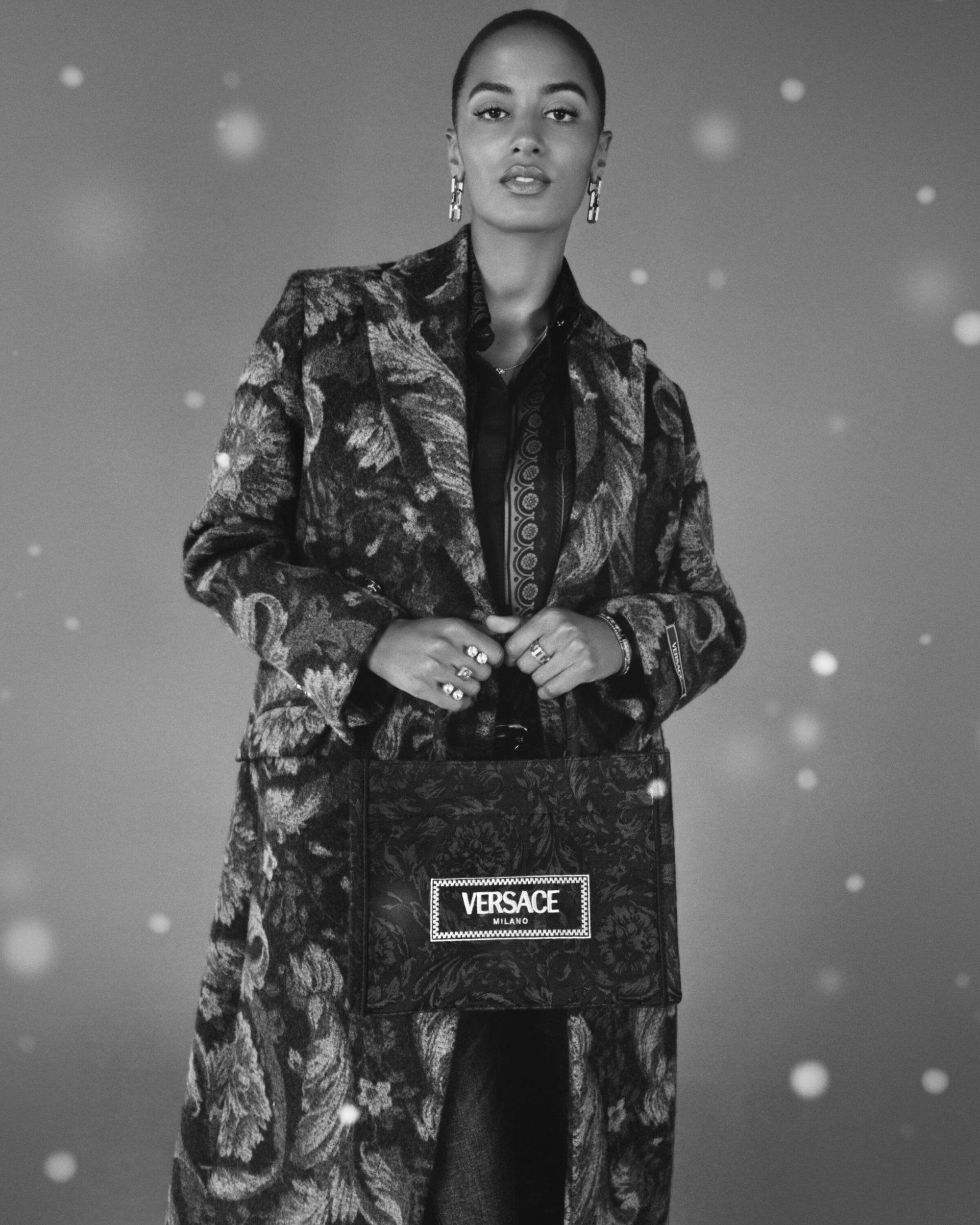 Black and white portrait of a woman in a patterned coat holding a Versace tote bag in a studio setting with subtle bokeh.