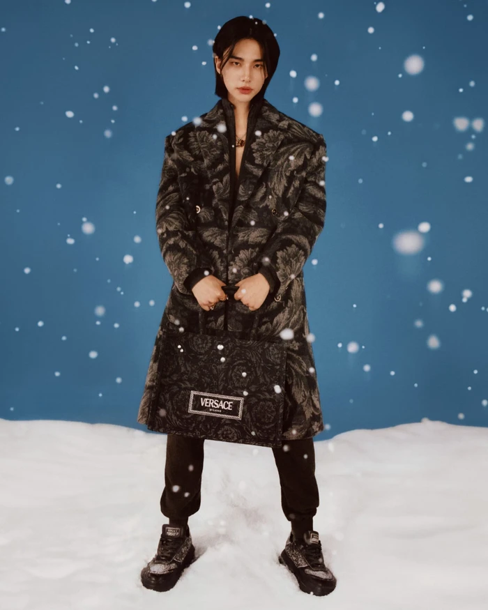 Hyunjin wearing a patterned black coat holding a matching Versace handbag in a snowy studio setting