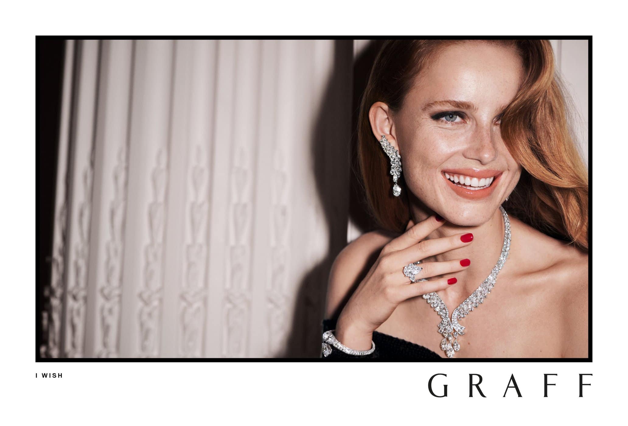 Smiling redhead woman showcasing Graff jewelry: diamond necklace, earrings, bracelet, and ring, against a draped fabric backdrop.