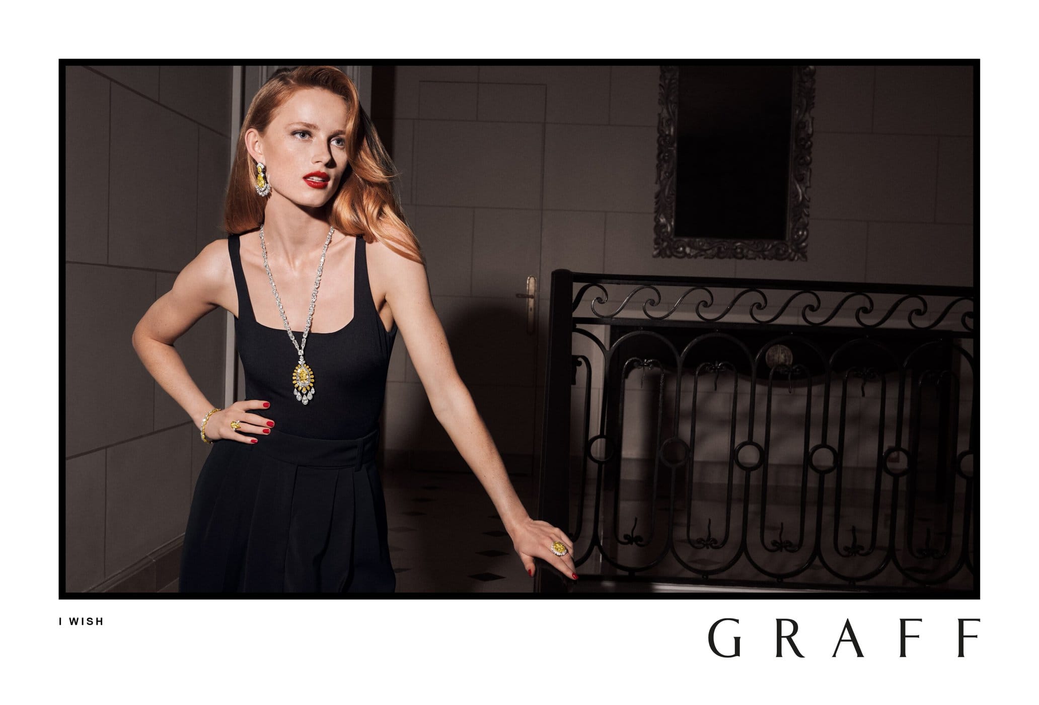 Elegant red-haired woman poses in a black dress and luxury jewelry in a high-fashion editorial photo, standing in an indoor setting.