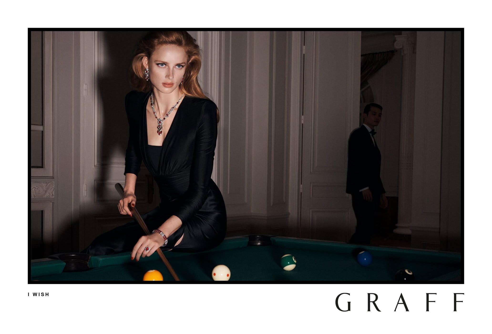 A woman in a black dress poses at a pool table with a man in a suit visible in the background, Graff jewelry, and a Graff logo.