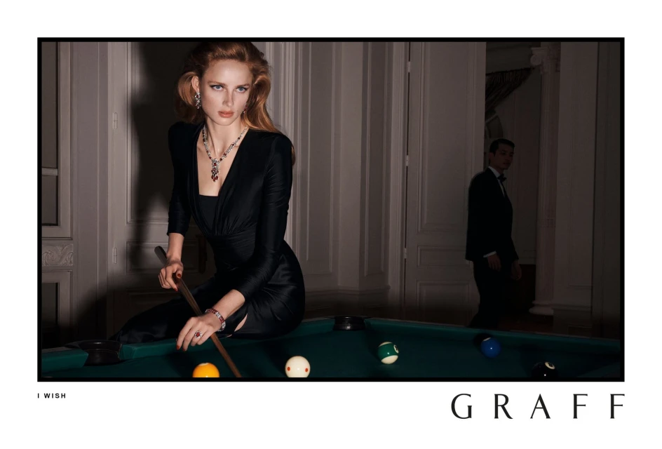 A woman in a black dress poses at a pool table with a man in a suit visible in the background, Graff jewelry, and a Graff logo.