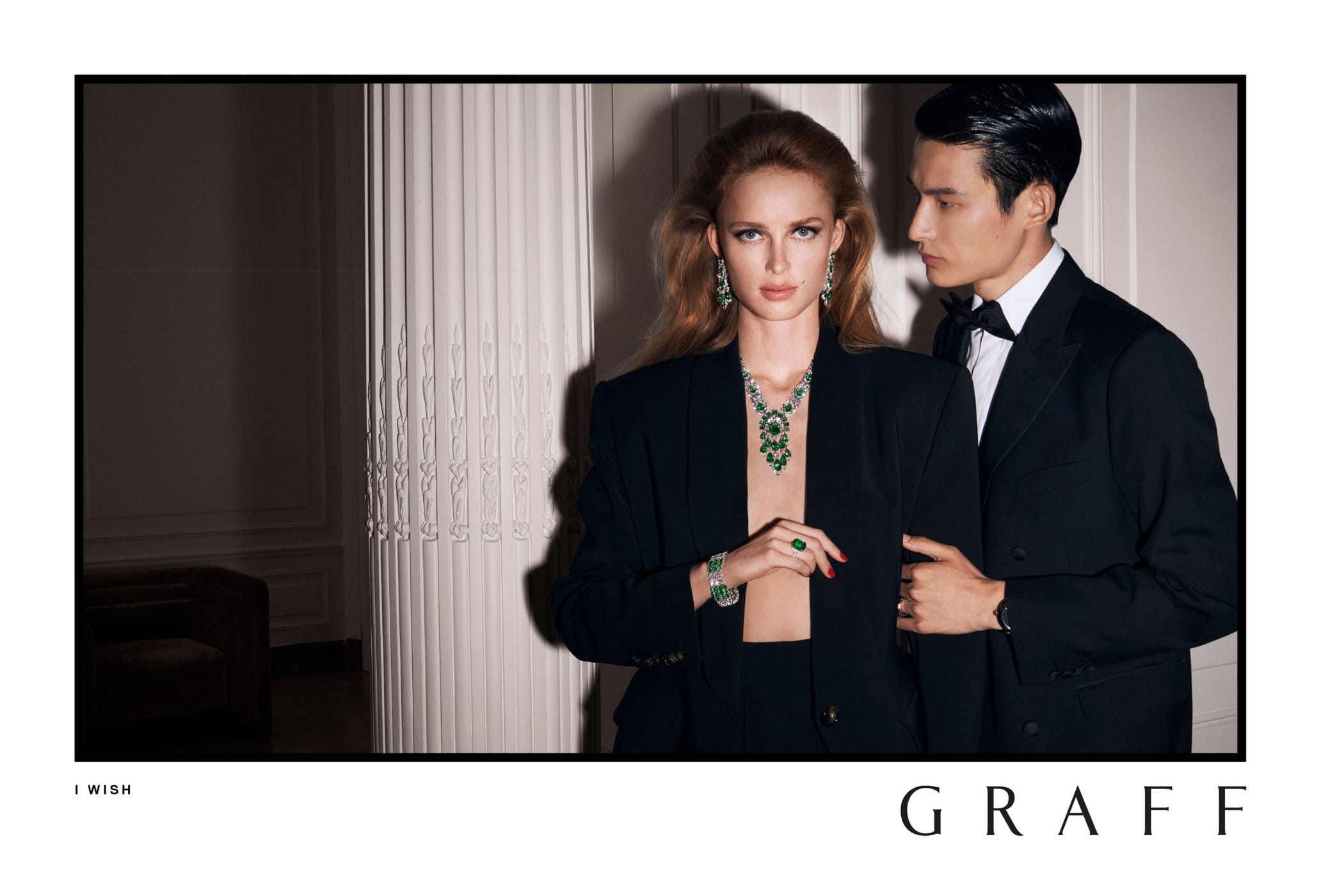 Model wearing emerald necklace and black blazer standing beside a man in a tuxedo