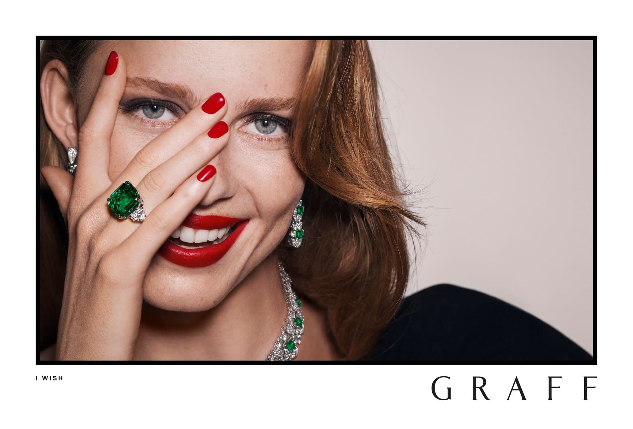 Portrait of a smiling adult female fashion model with red lipstick wearing emerald and diamond jewelry, against a neutral background.