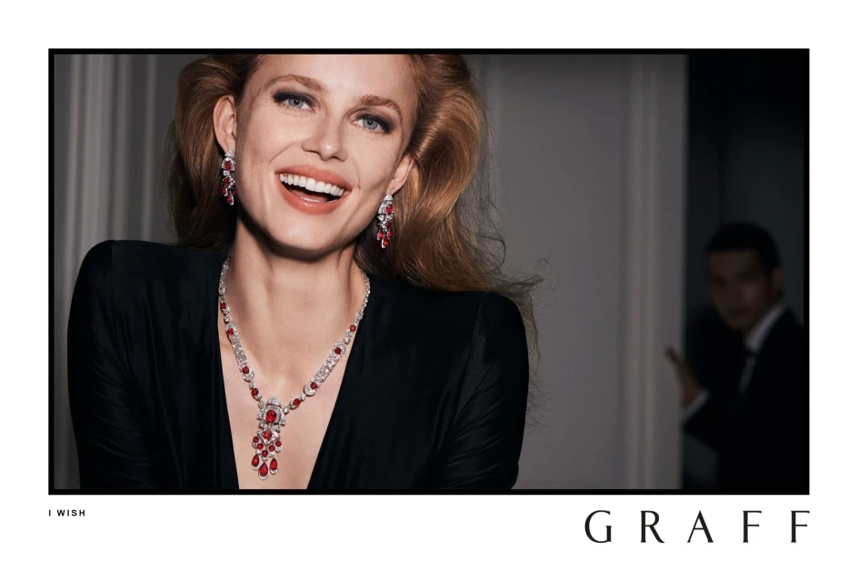 Close-up of a smiling woman with red hair wearing Graff jewelry: a necklace, earrings, and a black dress, set against a neutral background.