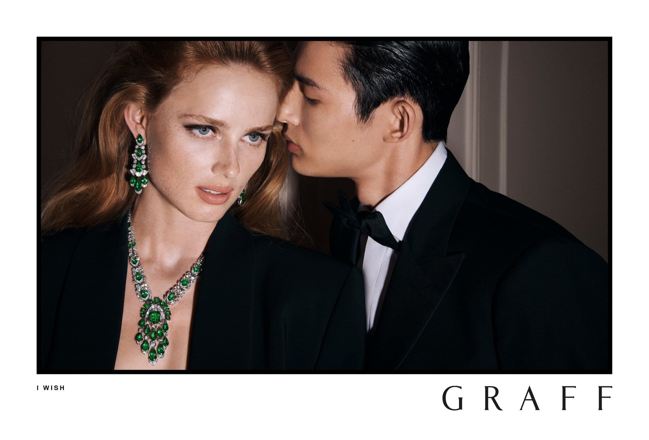 Portrait of a red-haired woman wearing green emerald jewelry, alongside an Asian man in a tuxedo, in a dimly lit indoor setting.