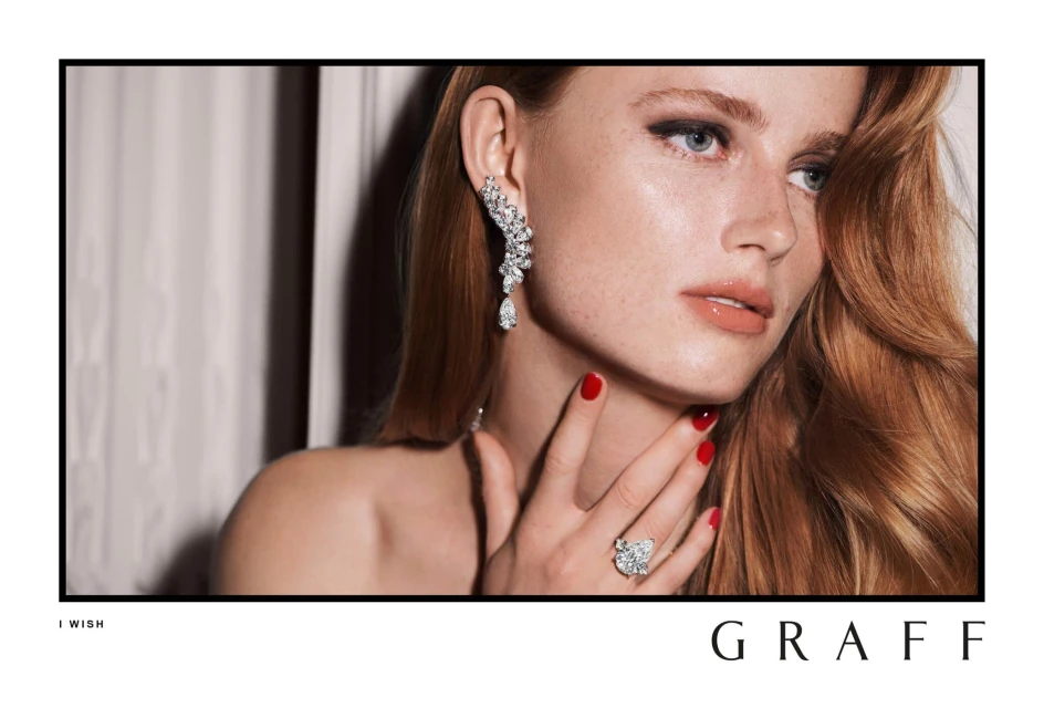 Portrait of a red-haired adult woman with red nails, wearing diamond jewelry, including an earring and ring, in a studio setting.