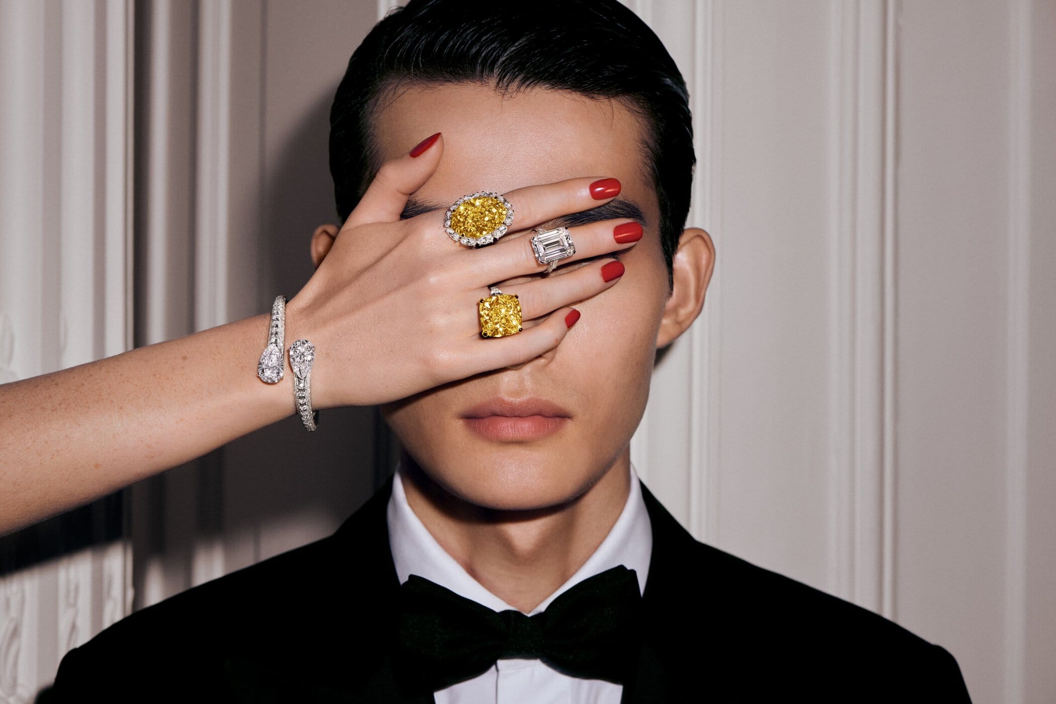 Close-up of a model in a suit with an outstretched hand, wearing diamond rings and a bracelet against a neutral background.