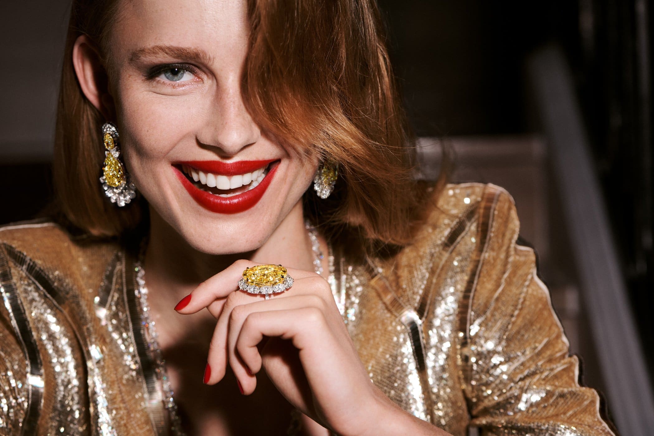 Smiling woman wearing a gold sequin jacket and a large yellow diamond ring, posed in a close-up beauty shot with red lipstick.