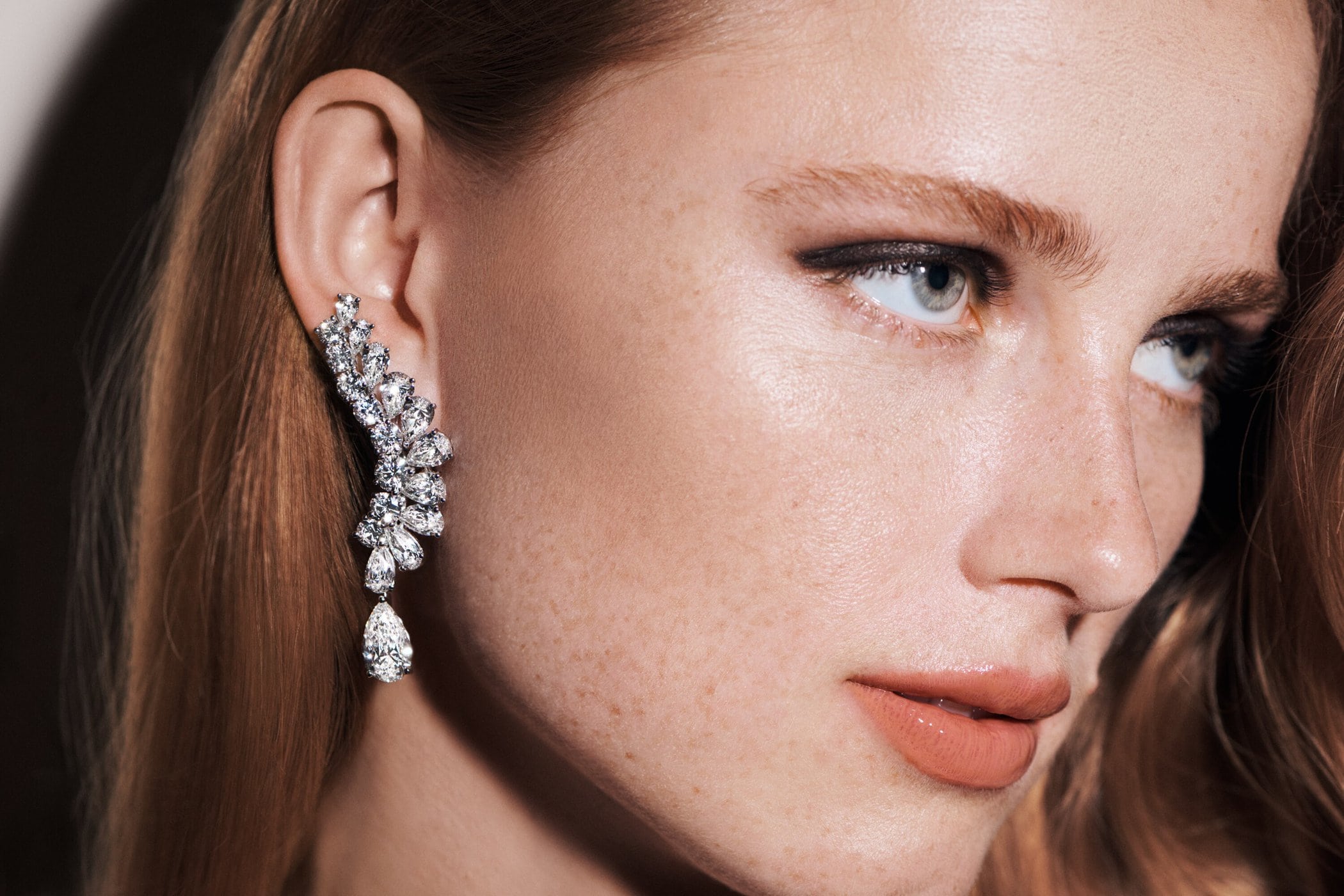 Close-up of a red-haired woman wearing an ornate, cascading diamond earring, showcasing beauty and fashion. Makeup highlights her eyes and lips.