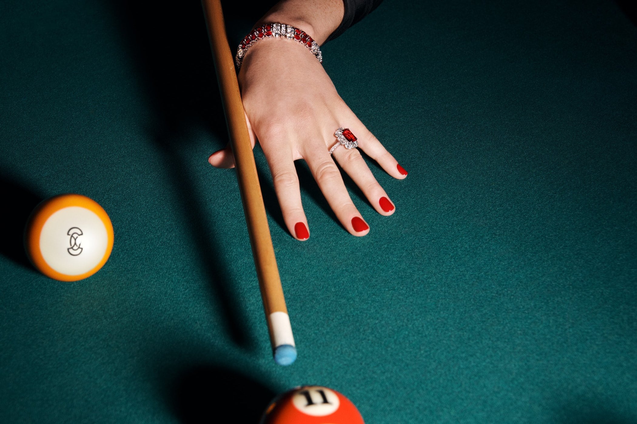 Elegant woman's hand with red manicure and jewelry holds a cue stick on a green felt pool table, with billiard balls.