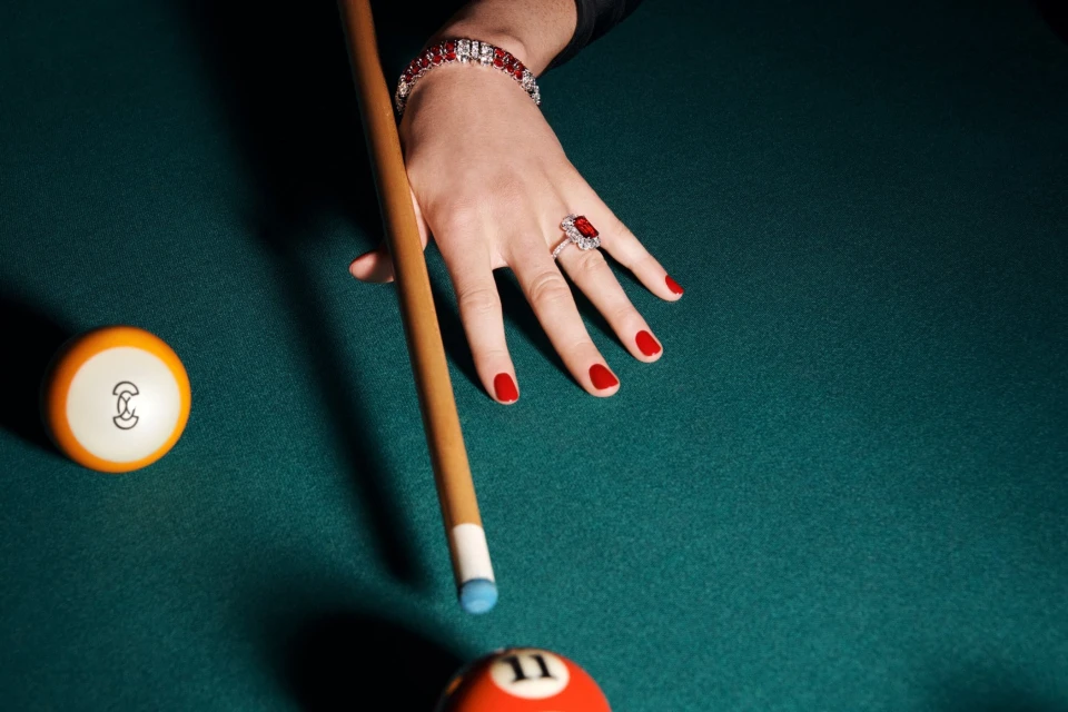 Elegant woman's hand with red manicure and jewelry holds a cue stick on a green felt pool table, with billiard balls.