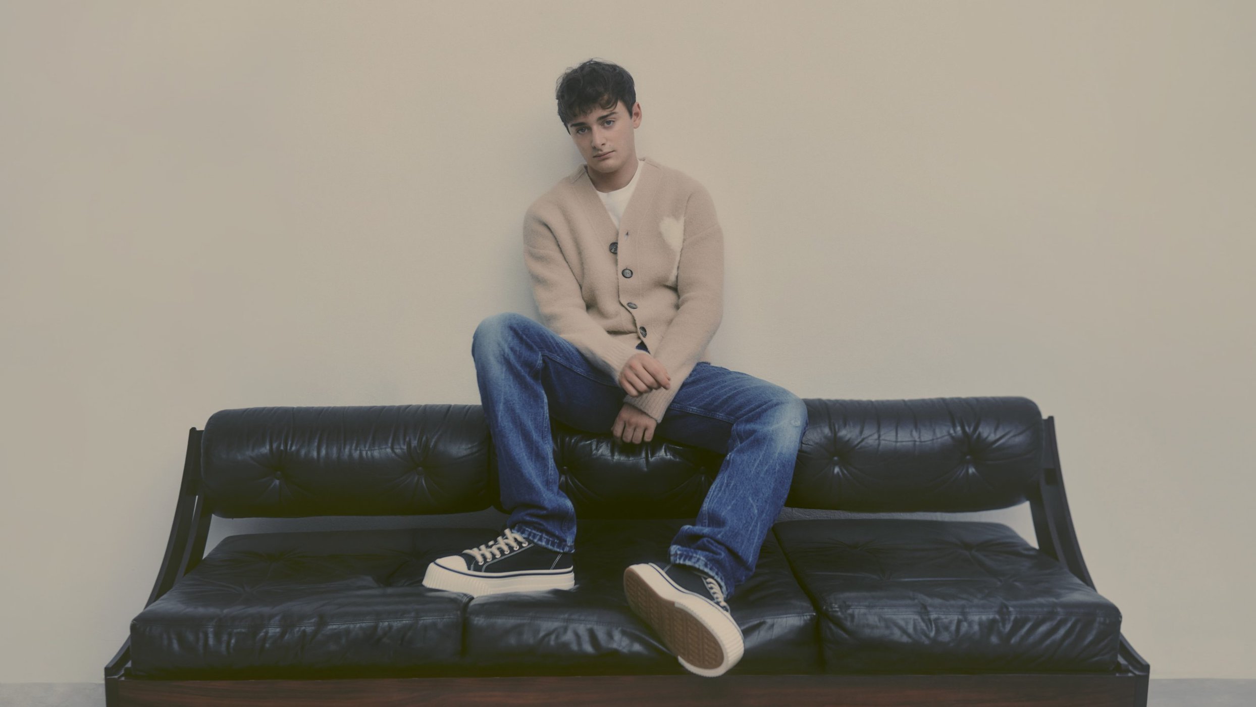 Adult male sitting on a leather couch, wearing a cardigan sweater and blue jeans, and black sneakers, against a neutral wall.