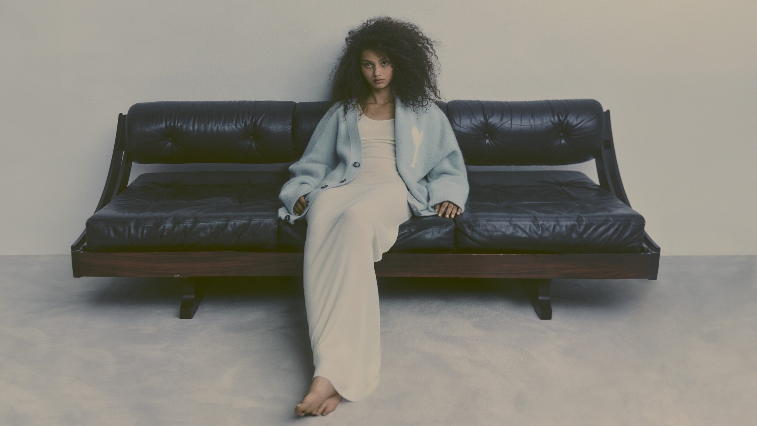 A young woman with curly hair sitting on a vintage black leather sofa wearing a long dress and cardigan in a minimalist studio setting.