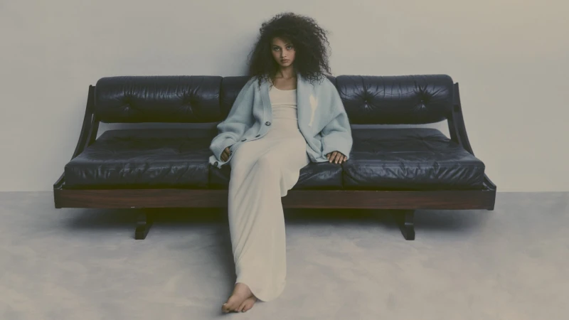 A young woman with curly hair sitting on a vintage black leather sofa wearing a long dress and cardigan in a minimalist studio setting.