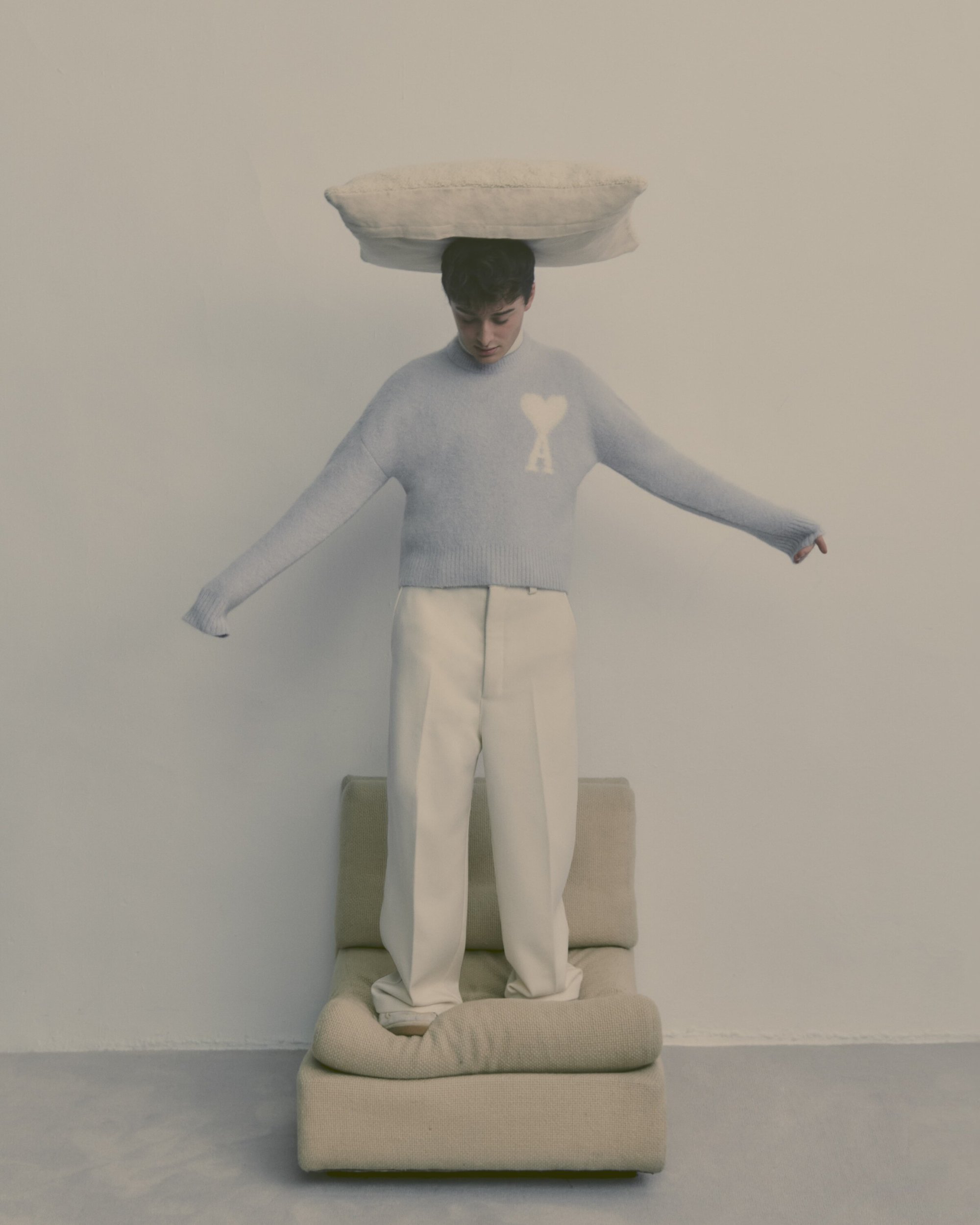 Man in a light blue sweater and white pants balances a pillow on his head, standing on a beige chair in a minimalist, light-toned setting.