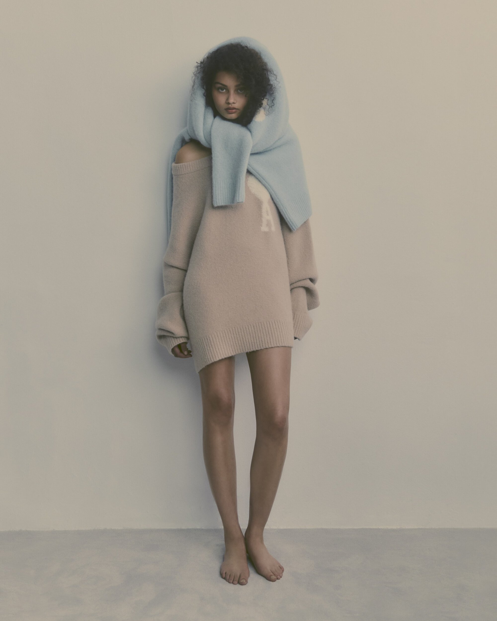 Woman in a beige sweater dress and light blue scarf, posing in a minimalist style in a neutral studio setting. Fashion editorial.
