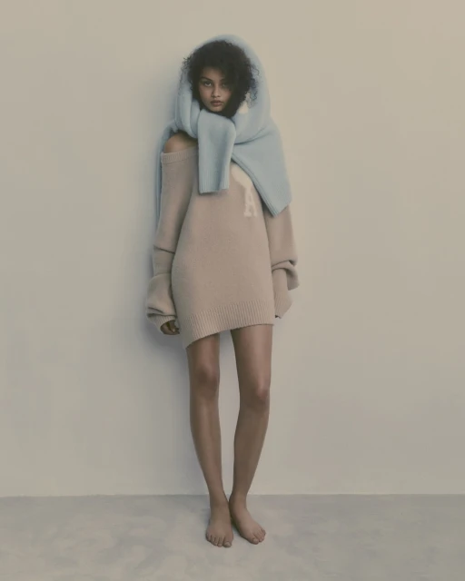 Woman in a beige sweater dress and light blue scarf, posing in a minimalist style in a neutral studio setting. Fashion editorial.