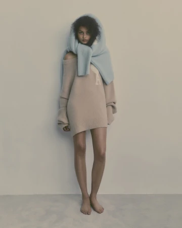 Woman in a beige sweater dress and light blue scarf, posing in a minimalist style in a neutral studio setting. Fashion editorial.