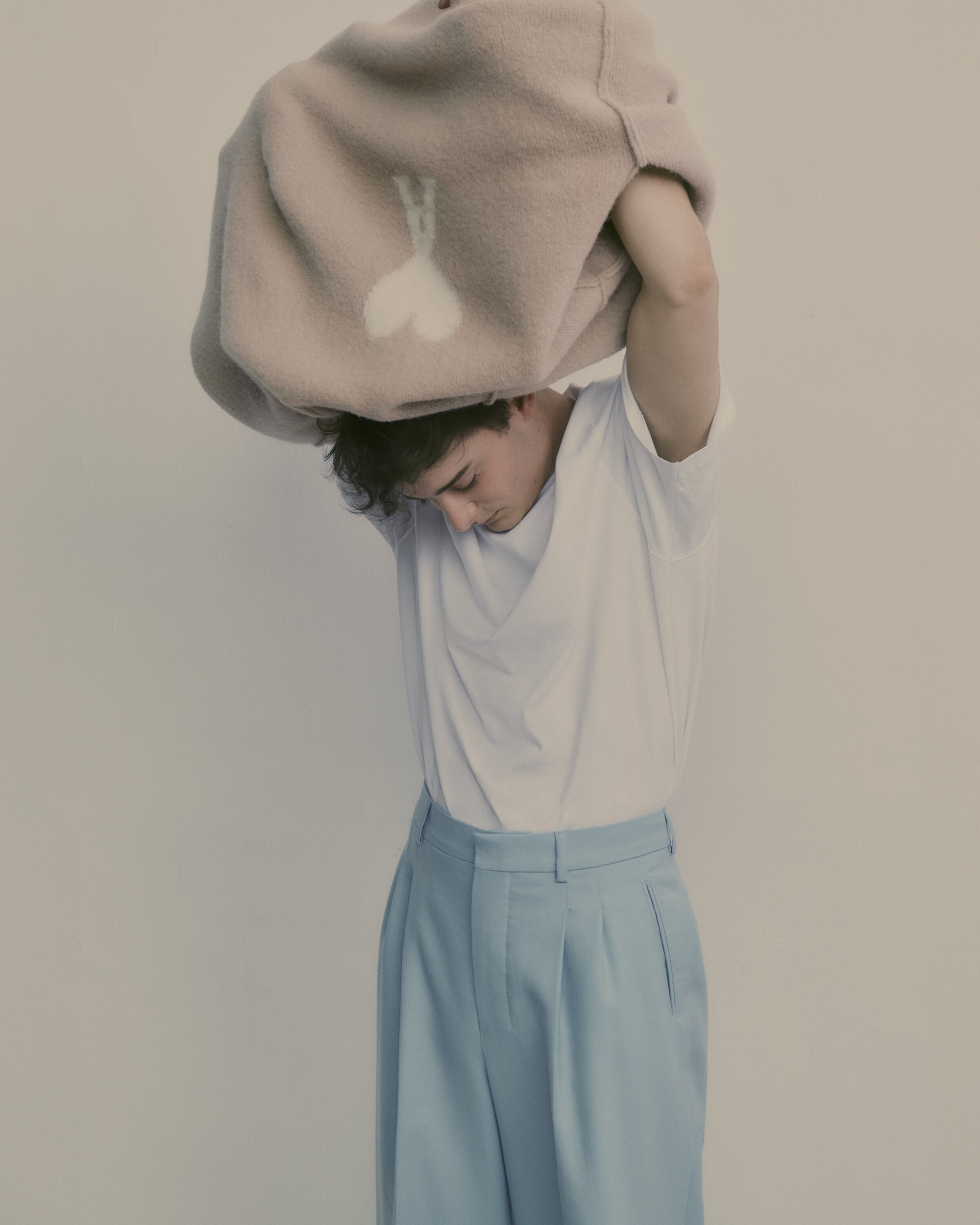 Young adult male is pictured indoors, putting a cream-colored sweater with white heart design over his head, wearing a white t-shirt and light blue trousers.