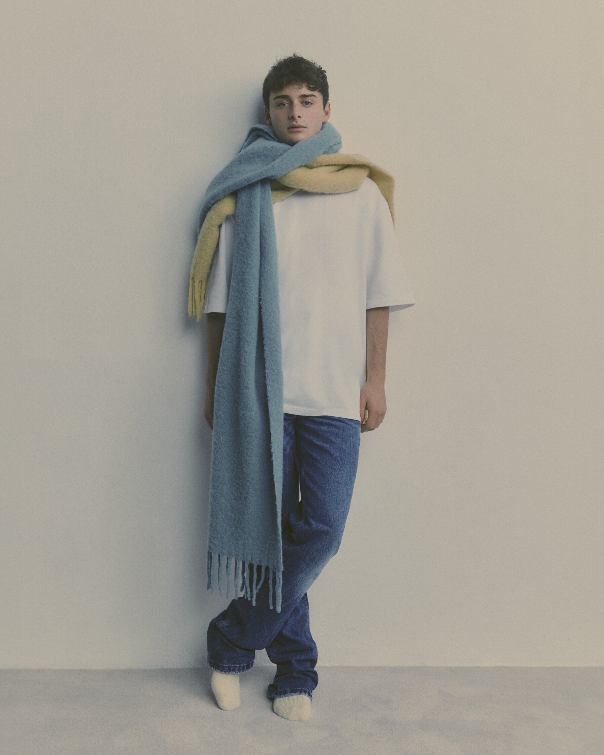Portrait of a young adult male wearing a white t-shirt, dark jeans, white socks, and a long blue and yellow scarf. Indoor studio setting.