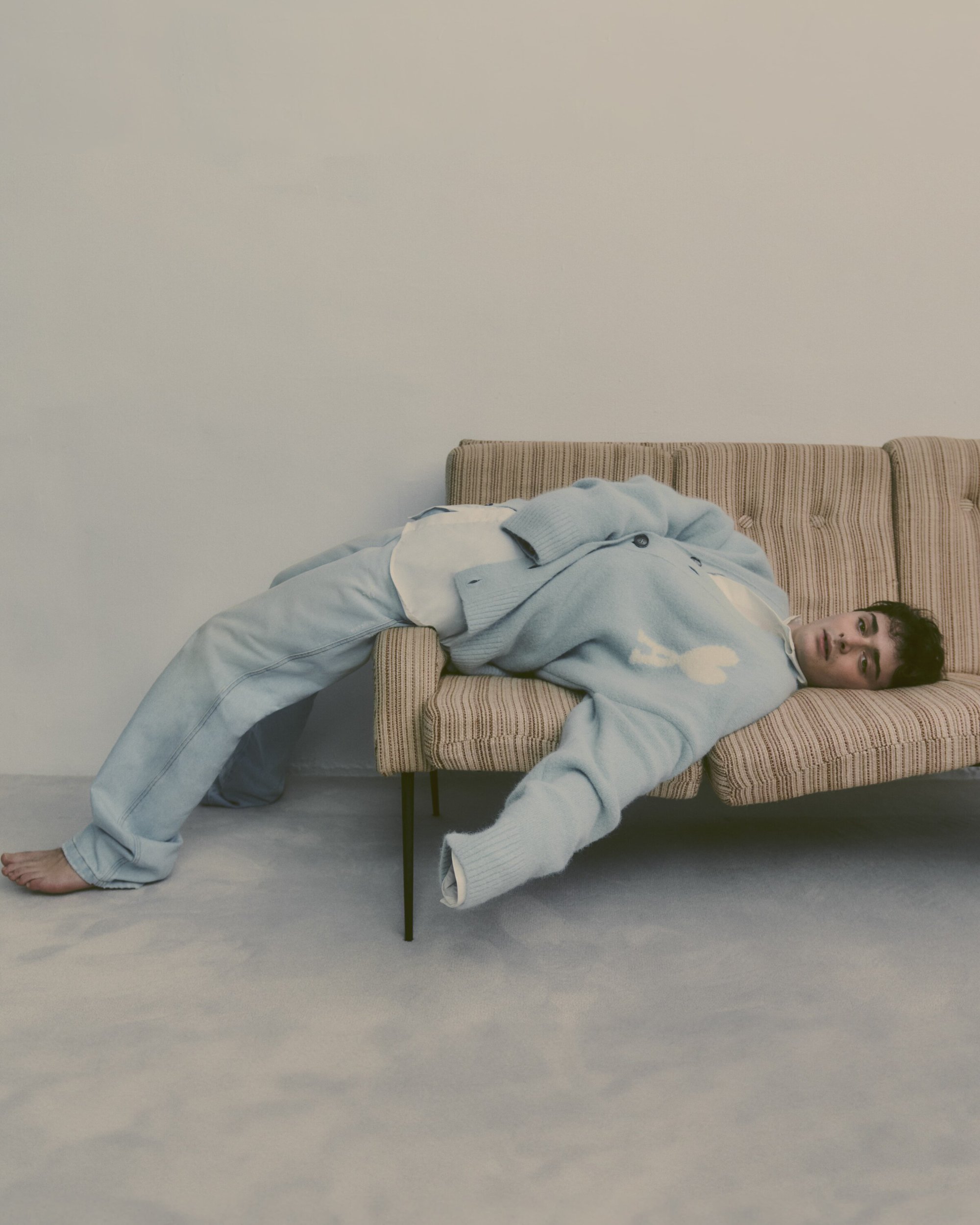 Adult male lounging on a striped couch in a photo studio. He is wearing a baby-blue cardigan and matching pants, creating a relaxed, casual look.