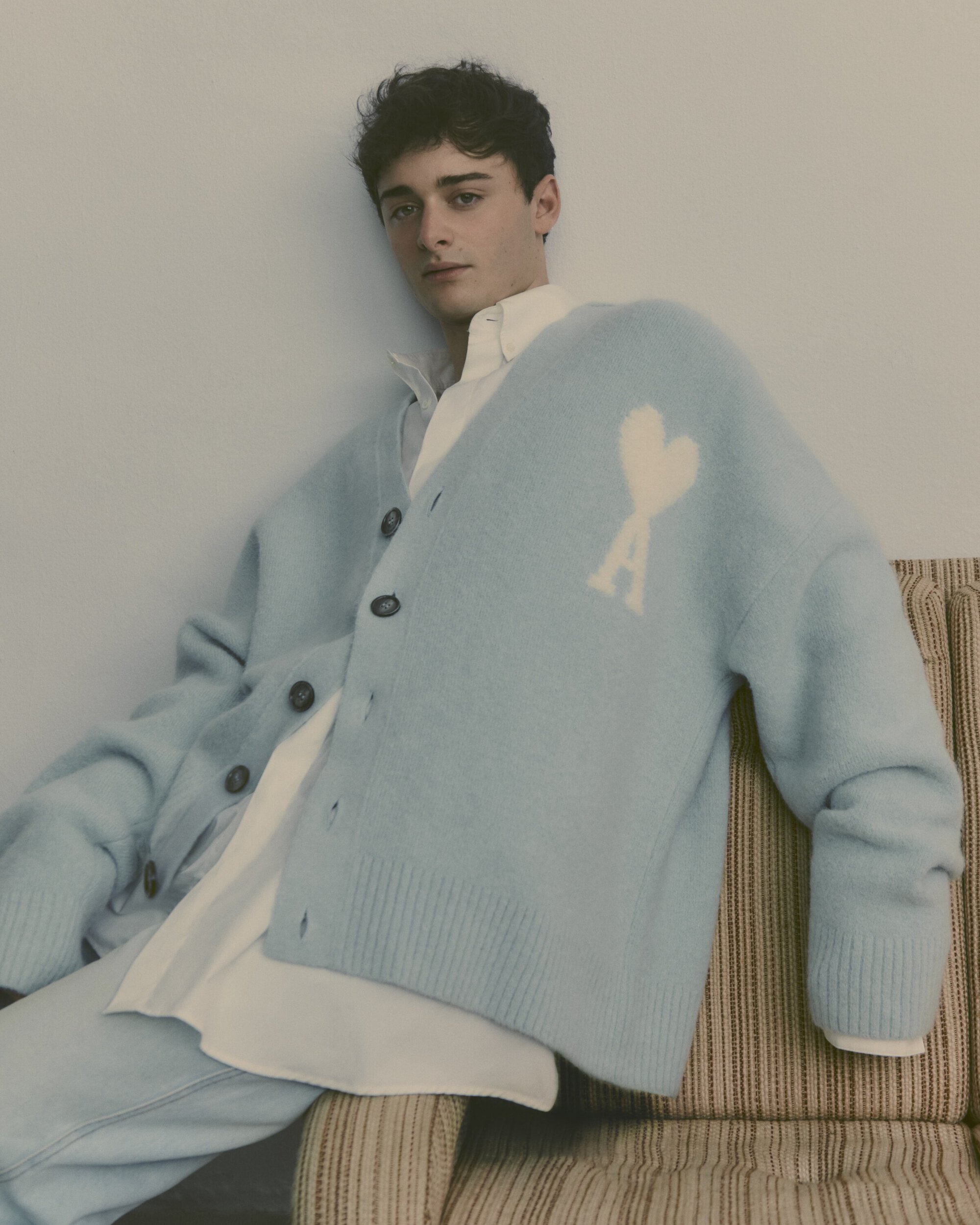 Adult male model poses indoors wearing a light blue cardigan sweater with a heart design and a white collared shirt.