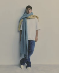 Noah Schnapp leaning against a plain grey studio wall wearing a white t-shirt, blue jeans, and draped wool scarves.