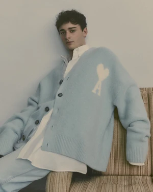 Noah Schnapp wearing an oversized light blue Ami cardigan and white shirt while seated in a striped vintage armchair.