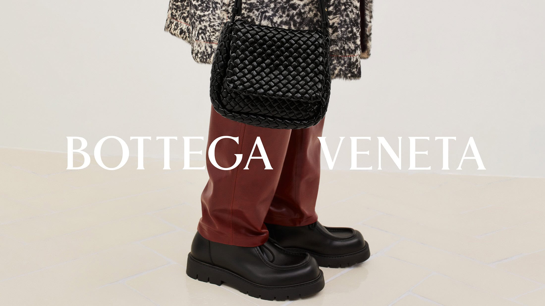 Fashion shot of a black woven handbag, black loafers, and maroon trousers on a person against a white background with brand logo.