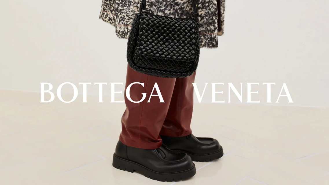 Fashion shot of a black woven handbag, black loafers, and maroon trousers on a person against a white background with brand logo.
