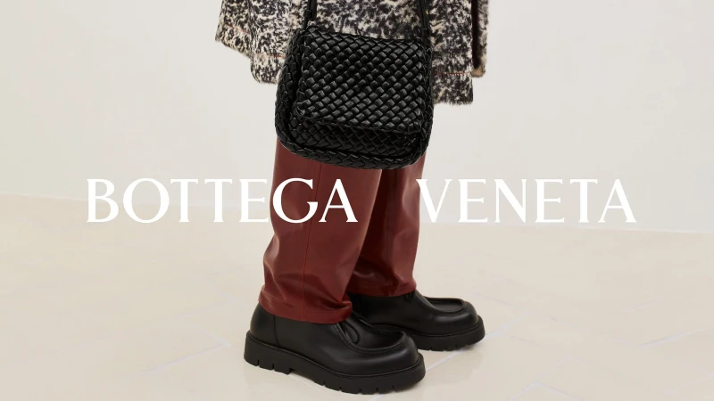Fashion shot of a black woven handbag, black loafers, and maroon trousers on a person against a white background with brand logo.