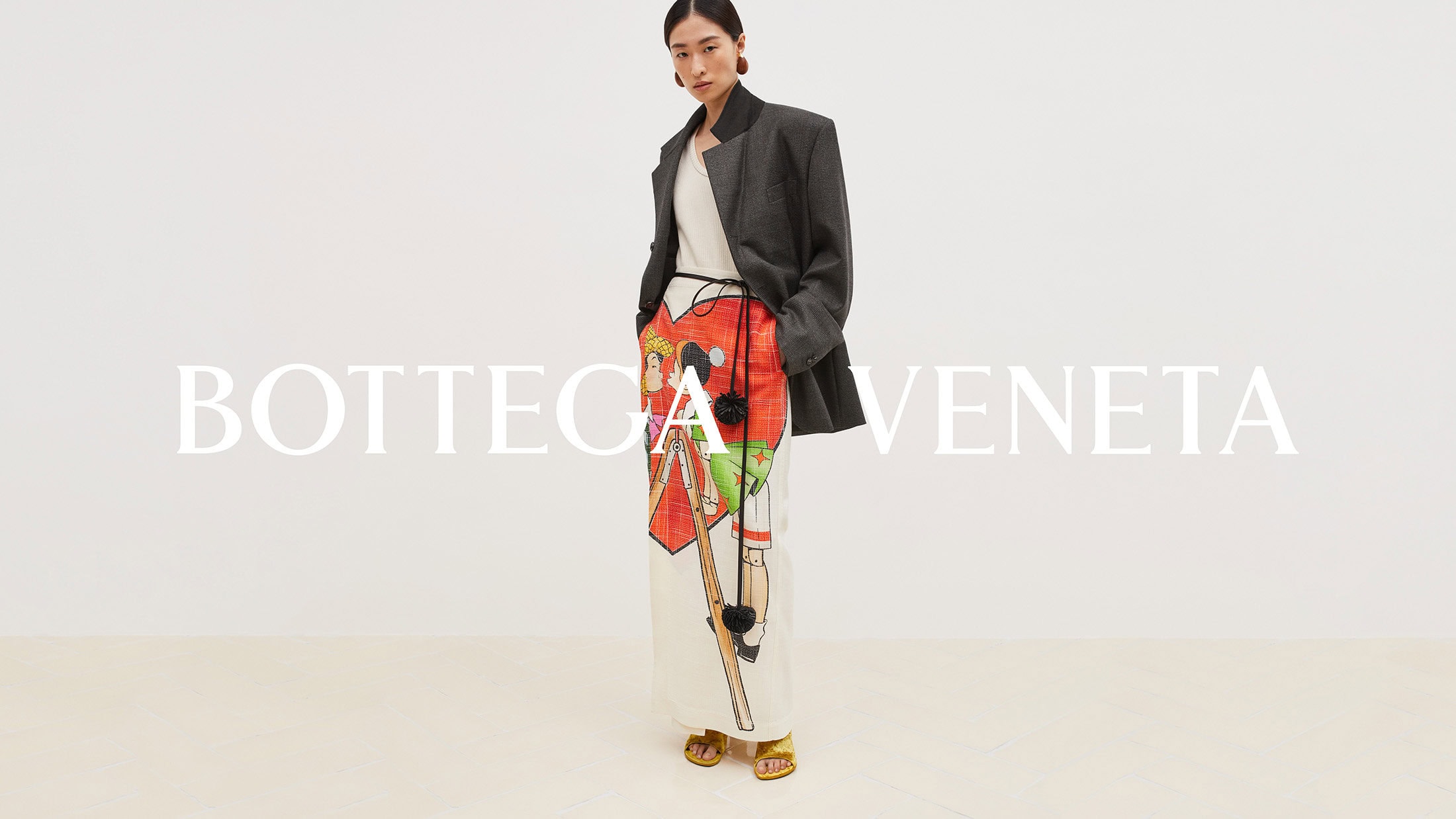 Fashion shot of a model wearing a grey blazer and illustrated white trousers with gold sandals, standing in front of the Bottega Veneta logo.