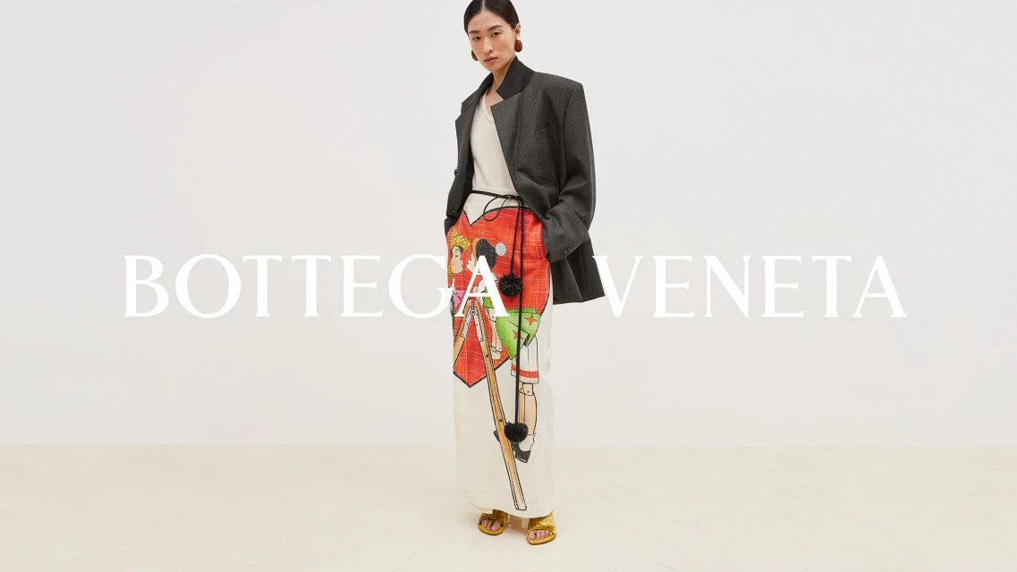 Fashion shot of a model wearing a grey blazer and illustrated white trousers with gold sandals, standing in front of the Bottega Veneta logo.
