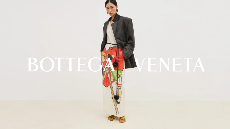 Fashion shot of a model wearing a grey blazer and illustrated white trousers with gold sandals, standing in front of the Bottega Veneta logo.