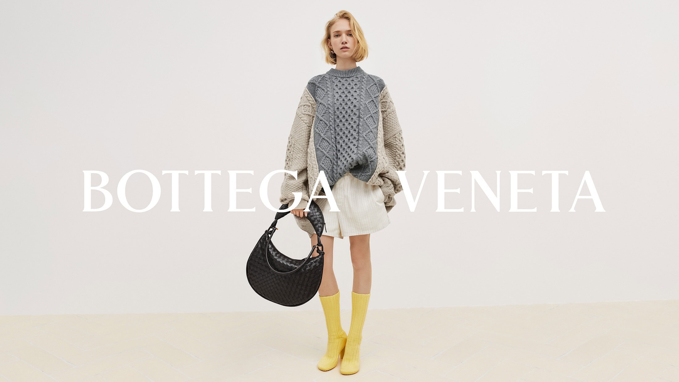 Fashion shoot of a woman wearing a patterned sweater, white shorts, and yellow boots, holding a Bottega Veneta purse against a white backdrop.