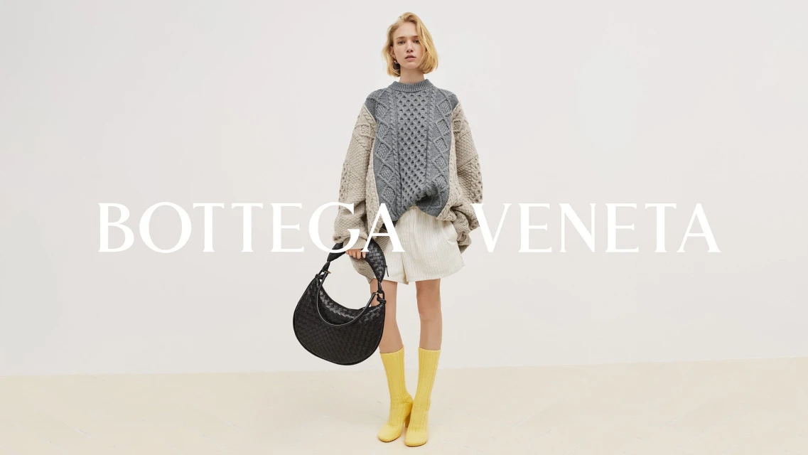 Fashion shoot of a woman wearing a patterned sweater, white shorts, and yellow boots, holding a Bottega Veneta purse against a white backdrop.